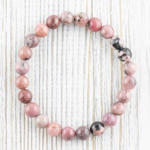 May include: A pink and black gemstone bracelet with round beads. The bracelet is made of natural stones and is strung on a clear elastic cord.