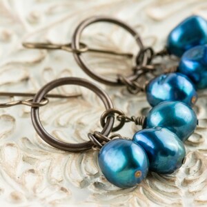 K&ouml;nnte beinhalten: Ein Paar Ohrringe mit dunkelbronzenen Reifen und Ohrhaken. Jeder Ohrring hat eine Ansammlung von f&uuml;nf irisierenden, tropfenf&ouml;rmigen blauen Perlen. Die Ohrringe liegen auf einer wei&szlig;en, verzierten Oberfl&auml;che.
