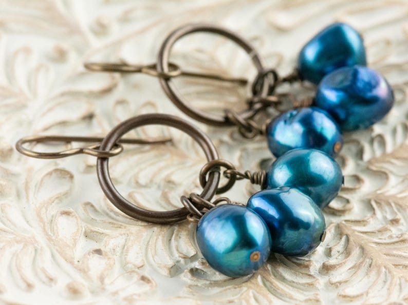 K&ouml;nnte beinhalten: Ein Paar Ohrringe mit dunkelbronzenen Reifen und Ohrhaken. Jeder Ohrring hat eine Ansammlung von f&uuml;nf irisierenden, tropfenf&ouml;rmigen blauen Perlen. Die Ohrringe liegen auf einer wei&szlig;en, verzierten Oberfl&auml;che.
