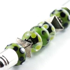 May include: A silver and green beaded bracelet with black and white polka dots. The bracelet has a silver cap on one end and a silver charm on the other.