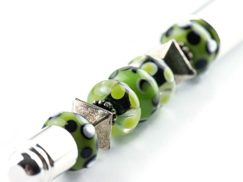 May include: A silver and green beaded bracelet with black and white polka dots. The bracelet has a silver cap on one end and a silver charm on the other.