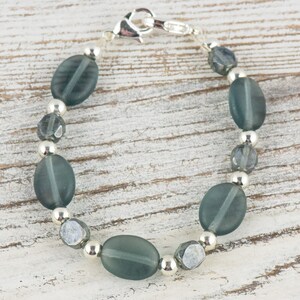 Pu&ograve; includere: Un braccialetto con perline ovali opache color teal, perline sfaccettate pi&ugrave; piccole e perline in metallo color argento. Il braccialetto &egrave; infilato su una catena color argento e ha una chiusura a moschettone. Il gioiello &egrave; esposto su una superficie di legno chiaro.