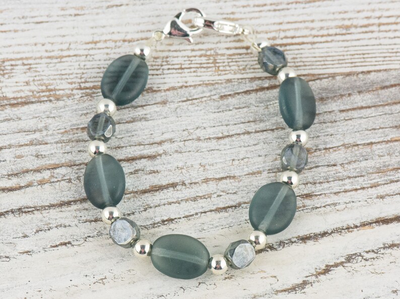 Pu&ograve; includere: Un braccialetto con perline ovali opache color teal, perline sfaccettate pi&ugrave; piccole e perline in metallo color argento. Il braccialetto &egrave; infilato su una catena color argento e ha una chiusura a moschettone. Il gioiello &egrave; esposto su una superficie di legno chiaro.