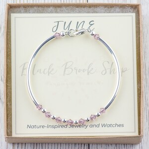 May include: A silver bangle bracelet with a small clasp and a row of light pink crystals. The bracelet is in a small box with a white card that says "JUNE" and "Black Brook Shop - Nature-Inspired Jewellery and Watches".