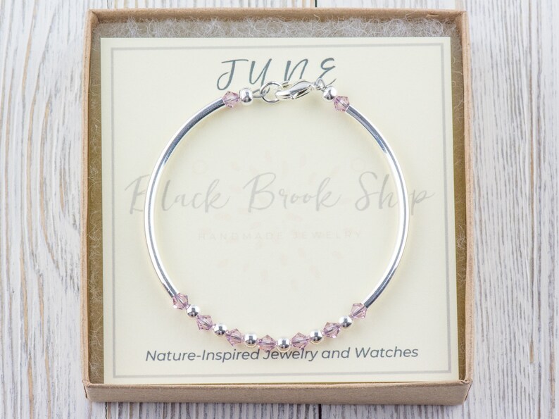 May include: A silver bangle bracelet with a small clasp and a row of light pink crystals. The bracelet is in a small box with a white card that says "JUNE" and "Black Brook Shop - Nature-Inspired Jewellery and Watches".