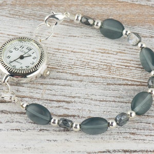 Pu&ograve; includere: Un bracciale orologio al quarzo Geneva argentato con quadrante bianco. Il bracciale presenta perline ovali grigie e perline argentate. Il quadrante mostra l'ora e la scritta "GENEVA".