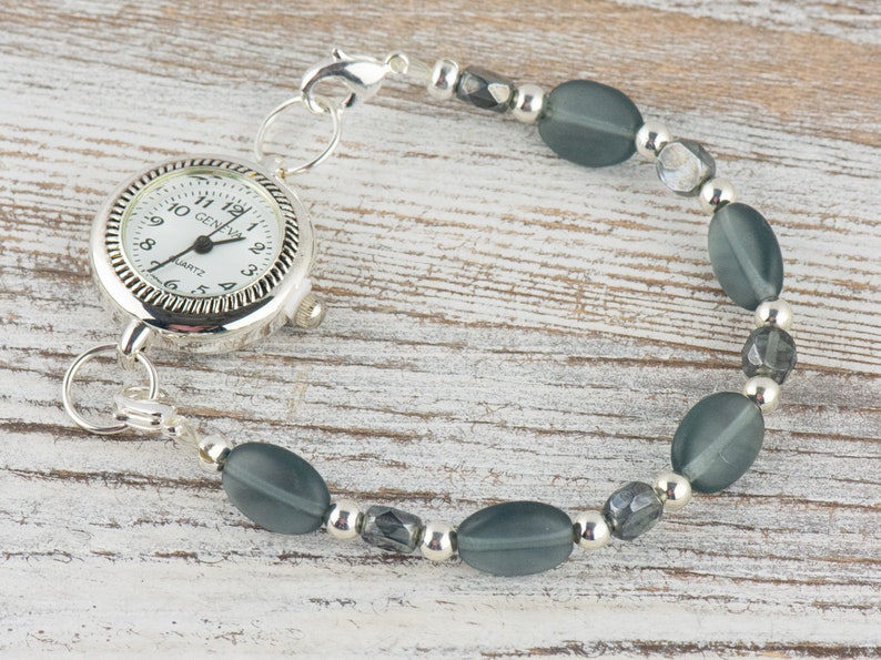 Pu&ograve; includere: Un bracciale orologio al quarzo Geneva argentato con quadrante bianco. Il bracciale presenta perline ovali grigie e perline argentate. Il quadrante mostra l'ora e la scritta "GENEVA".
