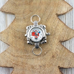 May include: Silver charm with a Santa Claus clock face. The clock face has black numbers and a white background. The charm has a silver loop for attaching to a bracelet or necklace.