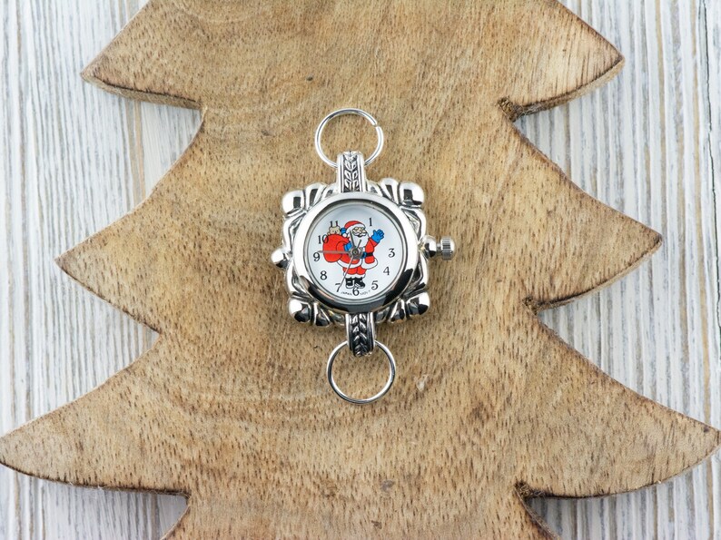 May include: Silver charm with a Santa Claus clock face. The clock face has black numbers and a white background. The charm has a silver loop for attaching to a bracelet or necklace.