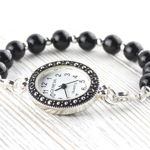 May include: A Geneva quartz watch bracelet with a white watch face and black numbers. The watch is surrounded by a silver-colored bezel and connected to a bracelet made of black, round beads. The watch is on a white wooden surface.