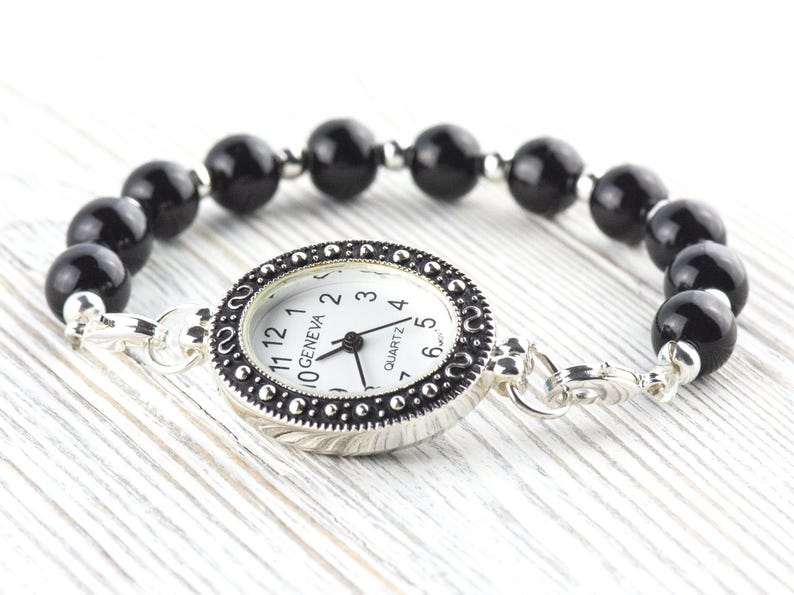 May include: A Geneva quartz watch bracelet with a white watch face and black numbers. The watch is surrounded by a silver-colored bezel and connected to a bracelet made of black, round beads. The watch is on a white wooden surface.