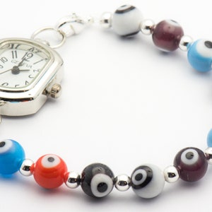 Puede incluir: Un reloj plateado con una esfera cuadrada y agujas negras est&aacute; unido a una pulsera con cuentas de color de ojo turco. Las cuentas son azules, rojas, negras, blancas y moradas.