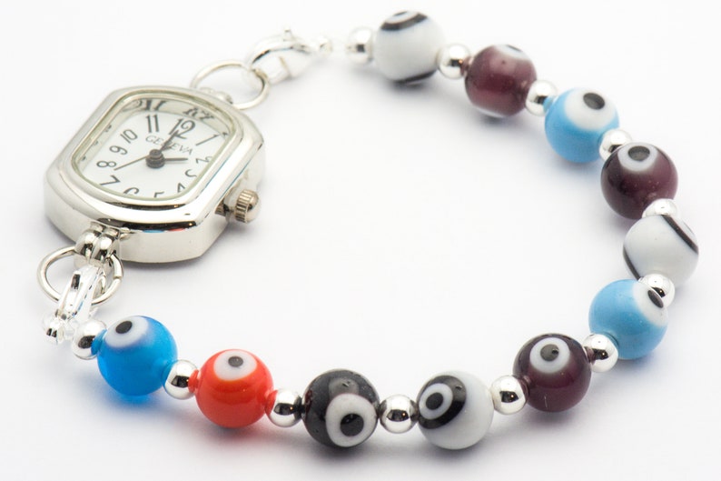 Puede incluir: Un reloj plateado con una esfera cuadrada y agujas negras est&aacute; unido a una pulsera con cuentas de color de ojo turco. Las cuentas son azules, rojas, negras, blancas y moradas.