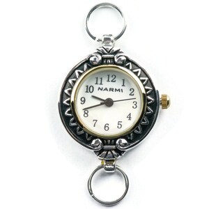 Pu&ograve; includere: Orologio da tasca argentato con quadrante bianco e dettagli dorati. L'orologio presenta una lunetta decorativa con un motivo e una piccola corona. Ha anelli argentati nella parte superiore e inferiore per il fissaggio.