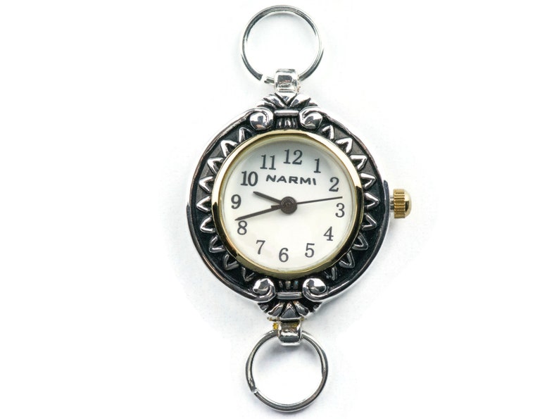 Pu&ograve; includere: Orologio da tasca argentato con quadrante bianco e dettagli dorati. L'orologio presenta una lunetta decorativa con un motivo e una piccola corona. Ha anelli argentati nella parte superiore e inferiore per il fissaggio.