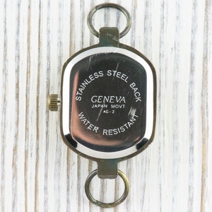 Pu&ograve; includere: Il retro di un orologio argentato con un quadrante quadrato. L'orologio ha il testo "STAINLESS STEEL BACK" e "GENEVA JAPAN MOVT AG-2" e "WATER RESISTANT" stampato sul retro.