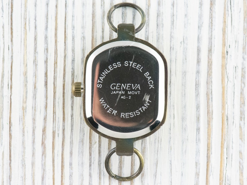 Pu&ograve; includere: Il retro di un orologio argentato con un quadrante quadrato. L'orologio ha il testo "STAINLESS STEEL BACK" e "GENEVA JAPAN MOVT AG-2" e "WATER RESISTANT" stampato sul retro.