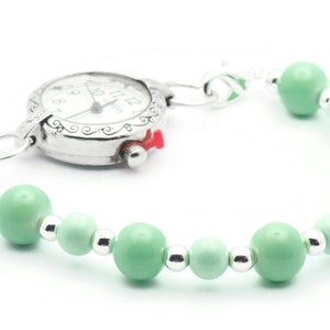 May include: A silver pocket watch with a white face and black hands attached to a green beaded bracelet.