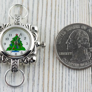 May include: Silver tone Christmas tree watch charm with a white face and black numbers. The watch charm is about the size of a UK 20 pence coin.