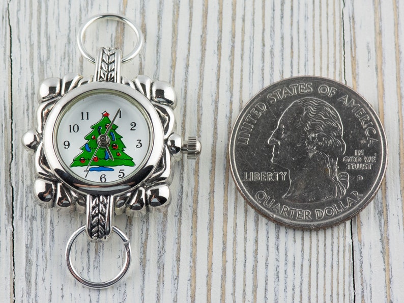 May include: Silver tone Christmas tree watch charm with a white face and black numbers. The watch charm is about the size of a UK 20 pence coin.