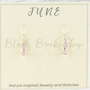 May include: A pair of dangle earrings with pink crystal beads and silver-tone metal accents. The earrings are displayed on a cream-colored card with the word "JUNE" and "Black Brook Shop" printed on it. The card also says "Nature-Inspired Jewelry and Watches."
