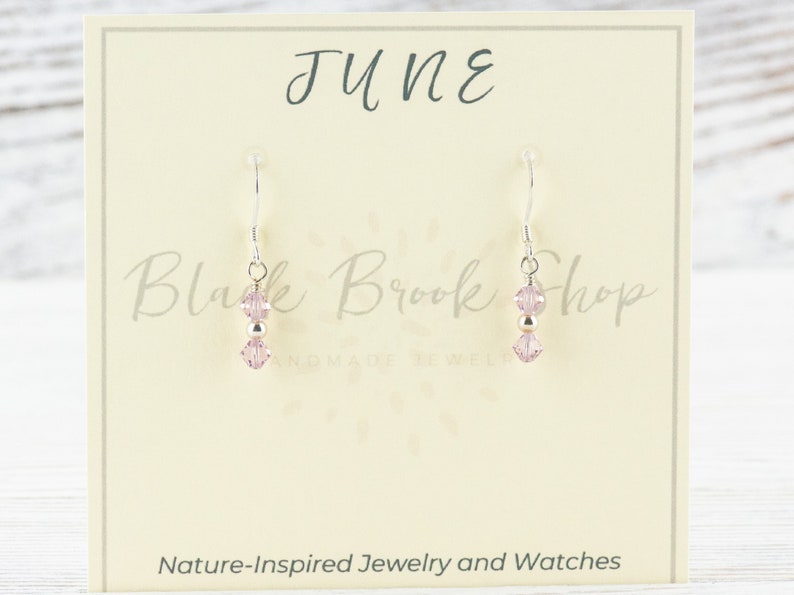 May include: A pair of dangle earrings with pink crystal beads and silver-tone metal accents. The earrings are displayed on a cream-colored card with the word "JUNE" and "Black Brook Shop" printed on it. The card also says "Nature-Inspired Jewelry and Watches."