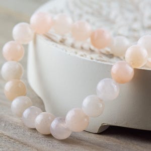 Op de afbeelding: Een armband gemaakt van lichtroze en witte kralen. De kralen zijn rond en glad en zijn geregen aan een doorzichtig elastiek.