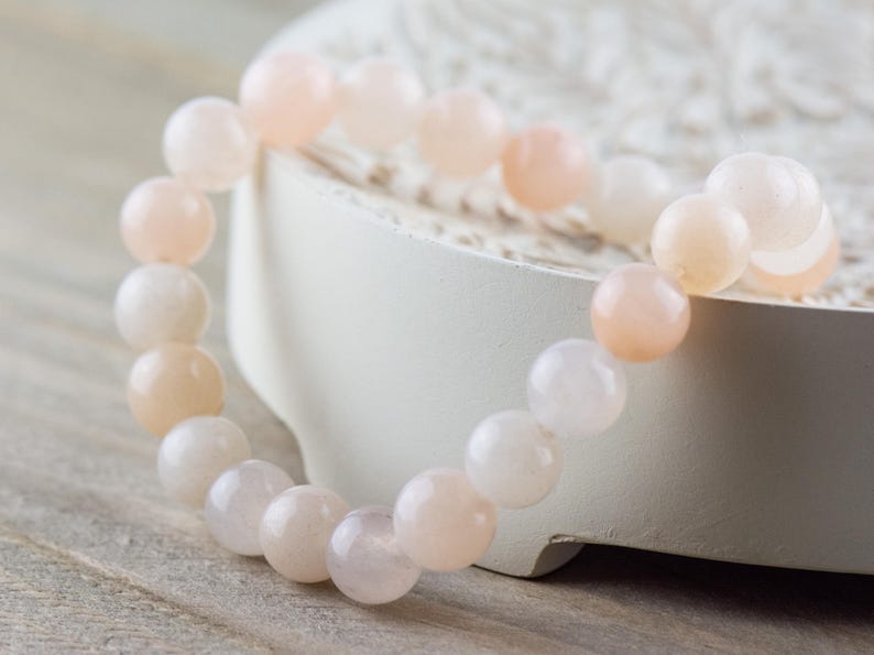Op de afbeelding: Een armband gemaakt van lichtroze en witte kralen. De kralen zijn rond en glad en zijn geregen aan een doorzichtig elastiek.