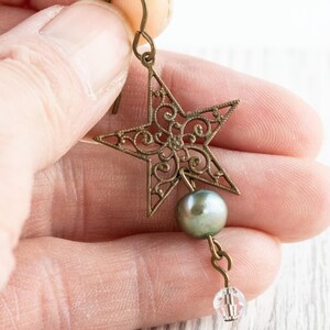 May include: A bronze-colored star-shaped earring with intricate filigree design. A gray pearl and a clear crystal bead dangle below the star. The earring is held up by a hook.