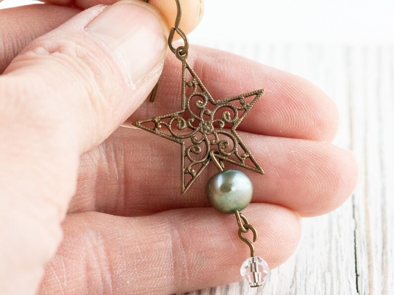 May include: A bronze-colored star-shaped earring with intricate filigree design. A gray pearl and a clear crystal bead dangle below the star. The earring is held up by a hook.