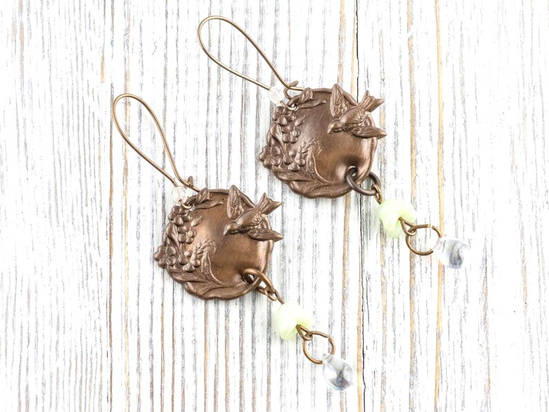 May include: A pair of bronze-colored earrings. Each earring features a circular pendant with a bird and grape design. The earrings have a long, curved hook and are adorned with a small, pale yellow bead and a clear, teardrop-shaped crystal.