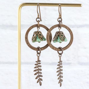 May include: A pair of handcrafted earrings with a botanical theme. Each earring features a bronze-coloured, textured hoop with a small, bell-shaped, light-green bead. A bronze fern-like charm dangles below the hoop, creating a nature-inspired design.