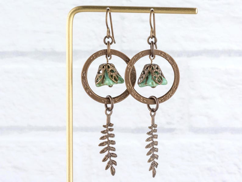May include: A pair of handcrafted earrings with a botanical theme. Each earring features a bronze-coloured, textured hoop with a small, bell-shaped, light-green bead. A bronze fern-like charm dangles below the hoop, creating a nature-inspired design.