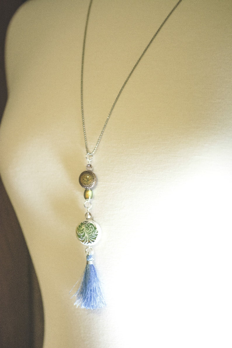 Puede incluir: Un collar de cadena plateada con un borla azul, una cuenta redonda con estampado verde y blanco y una cuenta redonda marr&oacute;n y dorada.