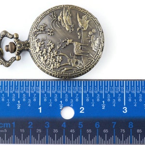 Pu&ograve; includere: Un orologio da tasca in bronzo con un design dettagliato di un cervo, alberi e montagne. L'orologio ha un diametro di 4,5 cm ed &egrave; dotato di una catena.