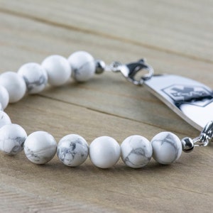 Peut inclure: Un bracelet en perles d'howlite blanches avec une plaque d'identification m&eacute;dicale en argent. La plaque porte un symbole m&eacute;dical et le mot "MedicAlert" grav&eacute;.