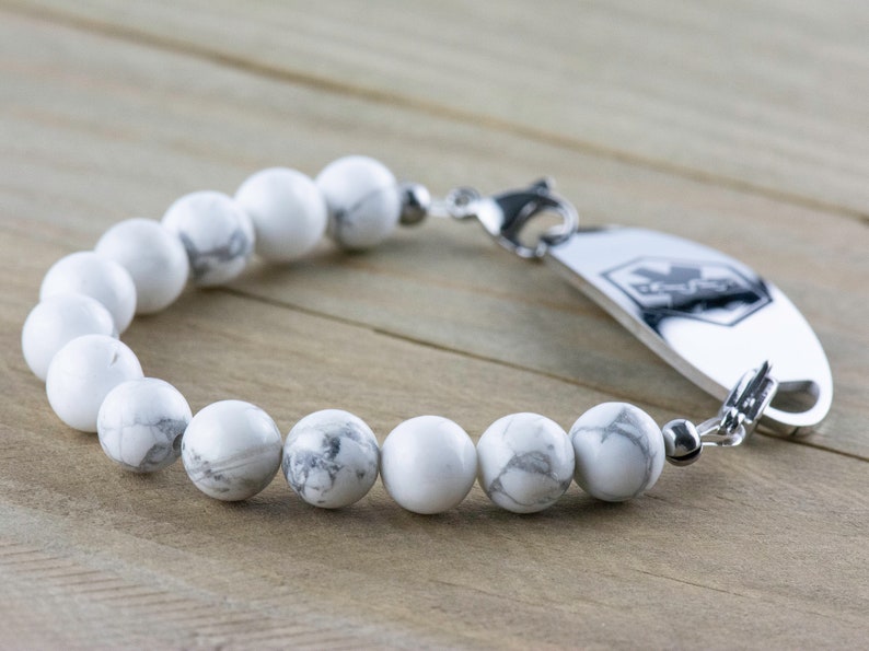 Peut inclure: Un bracelet en perles d'howlite blanches avec une plaque d'identification m&eacute;dicale en argent. La plaque porte un symbole m&eacute;dical et le mot "MedicAlert" grav&eacute;.