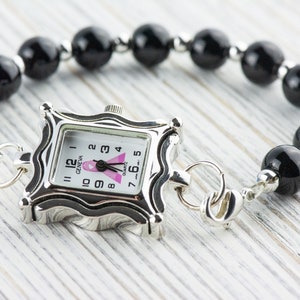 K&ouml;nnte beinhalten: Eine silberfarbene Quarzuhr mit quadratischem Zifferblatt und einem Armband aus schwarzen Perlen. Das Zifferblatt hat ein rosa Band und das Wort "GENEVA". Das Armband besteht aus gro&szlig;en, runden, schwarzen Perlen und silberfarbenen Akzenten.