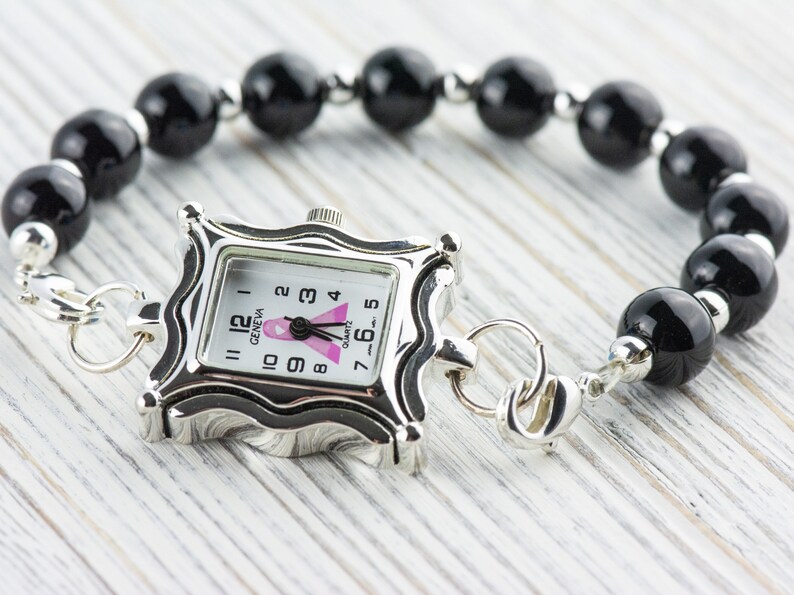 K&ouml;nnte beinhalten: Eine silberfarbene Quarzuhr mit quadratischem Zifferblatt und einem Armband aus schwarzen Perlen. Das Zifferblatt hat ein rosa Band und das Wort "GENEVA". Das Armband besteht aus gro&szlig;en, runden, schwarzen Perlen und silberfarbenen Akzenten.