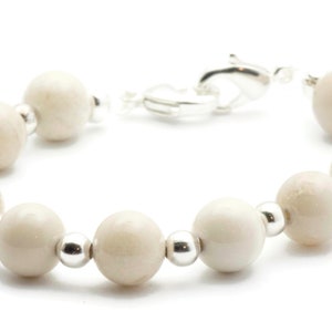 Op de afbeelding: Een witte kralen armband met zilveren accenten. De armband is gemaakt van ronde, gladde kralen en heeft een zilveren sluiting.