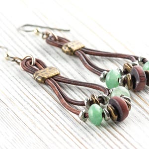 Pu&ograve; includere: Un paio di orecchini fatti a mano con cordoncini in pelle marrone, dettagli in metallo color ottone e perline verdi e marroni. Gli orecchini hanno uno stile boh&eacute;mien e sono esposti su una superficie di legno bianco.