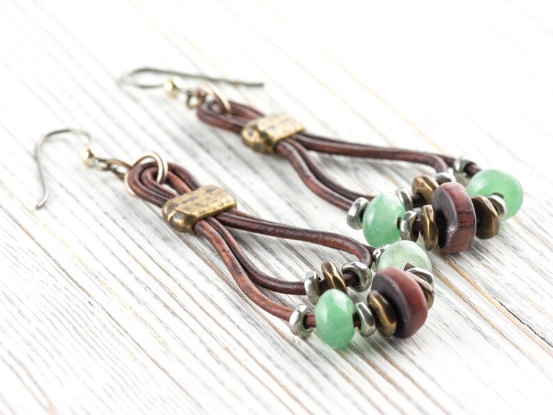 Pu&ograve; includere: Un paio di orecchini fatti a mano con cordoncini in pelle marrone, dettagli in metallo color ottone e perline verdi e marroni. Gli orecchini hanno uno stile boh&eacute;mien e sono esposti su una superficie di legno bianco.