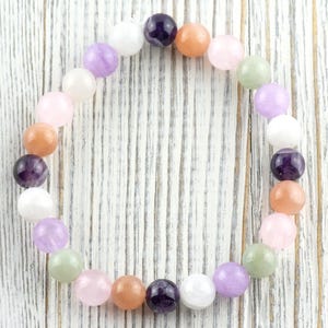 May include: A beaded bracelet featuring a variety of colorful gemstones. The bracelet includes round beads in shades of purple, pink, orange, white, and green. The bracelet is displayed on a white wooden surface.