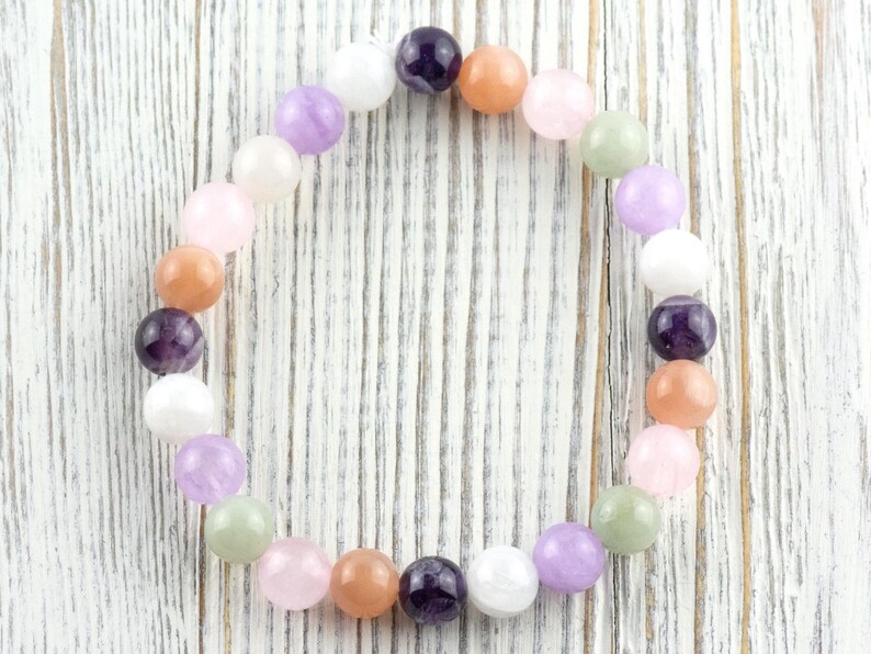 May include: A beaded bracelet featuring a variety of colorful gemstones. The bracelet includes round beads in shades of purple, pink, orange, white, and green. The bracelet is displayed on a white wooden surface.