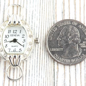May include: A silver-toned quartz watch with a white face and black numerals. The watch is oval-shaped and adorned with small, clear crystals. The watch face reads "GENEVA" and "QUARTZ". A U.S. quarter is next to the watch for size comparison.