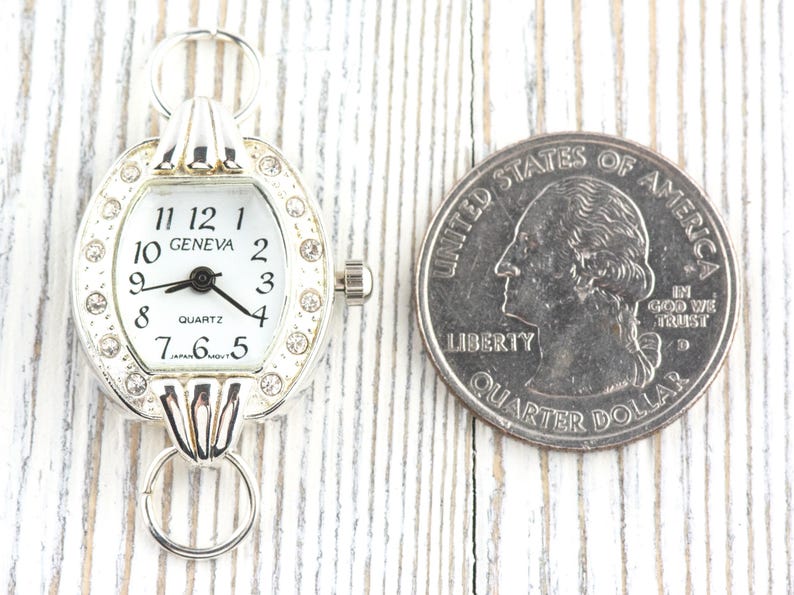 May include: A silver-toned quartz watch with a white face and black numerals. The watch is oval-shaped and adorned with small, clear crystals. The watch face reads "GENEVA" and "QUARTZ". A U.S. quarter is next to the watch for size comparison.