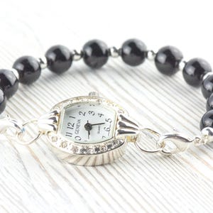 May include: A Geneva quartz watch bracelet featuring a silver-toned watch face with a white dial and black numbers. The bracelet is made of black, glossy, round beads. The watch is connected to the bracelet with silver-toned links.