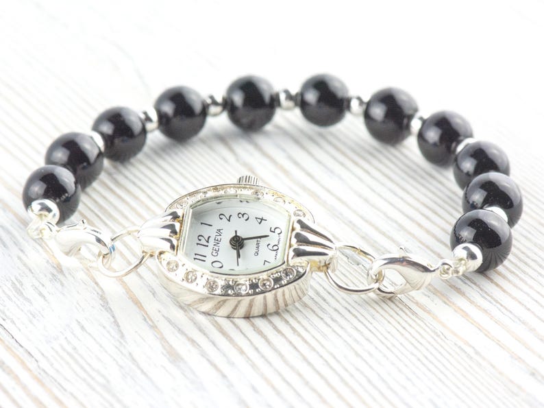 May include: A Geneva quartz watch bracelet featuring a silver-toned watch face with a white dial and black numbers. The bracelet is made of black, glossy, round beads. The watch is connected to the bracelet with silver-toned links.