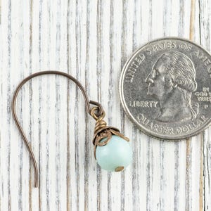 May include: A single earring with a light blue bead and bronze-colored metal accents. The earring is next to a U.S. quarter for size comparison. The background is a white-painted wooden surface.
