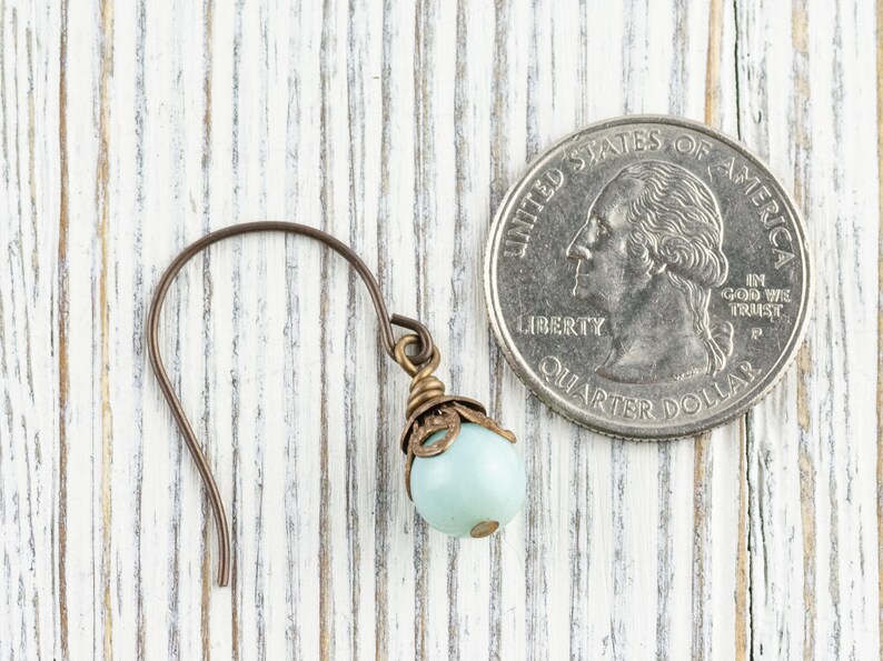 May include: A single earring with a light blue bead and bronze-colored metal accents. The earring is next to a U.S. quarter for size comparison. The background is a white-painted wooden surface.